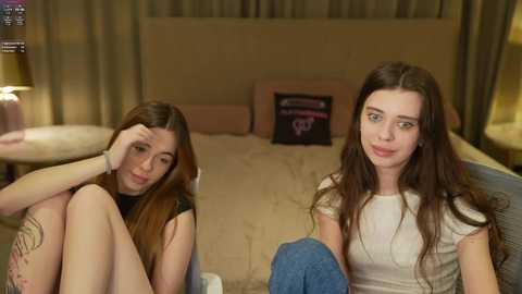 Video of two young women with long brown hair, one resting her head on her hand, the other smiling, in a cozy bedroom with beige walls and a headboard.