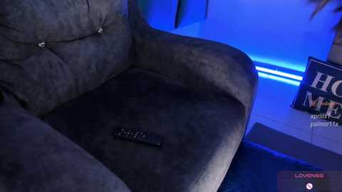 A video of a plush, dark grey couch with a remote control on the cushion, under blue LED lighting, with a wall-mounted \"Home\" sign in the background.