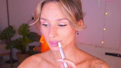 Video of a topless woman with light skin, blonde hair, and freckles, applying lipstick in a cozy room with string lights, plants, and a lamp.