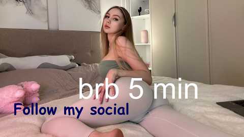 Video of a blonde woman with fair skin, wearing a strapless green top and white leggings, posing on a bed with gray and pink pillows, in a cozy bedroom. Text overlay reads \"brr 5 min show my social.\