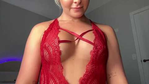 Video of a light-skinned woman with blonde hair, wearing a revealing red lace lingerie bodysuit with crisscross straps, smiling subtly. Background shows a plain, white-walled room with a door.