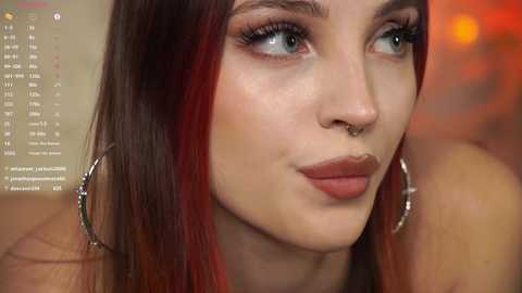 Video of a young woman with fair skin, red and black hair, light makeup, and hoop earrings, smiling with a septum ring, in a blurred, warm-toned background.