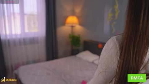 Video of a woman with long brown hair sitting on a bed with white linens, looking towards a softly lit room with a potted plant and a lamp.