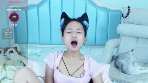 Video of an Asian woman with black cat ears, wearing a pink top, lying on a bed, eyes closed, mouth open, surrounded by stuffed animals and a cat.