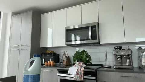 Video of a modern kitchen with sleek white cabinets, stainless steel appliances, a microwave, toaster oven, and a coffee maker. Bright white subway tiles cover the backsplash.