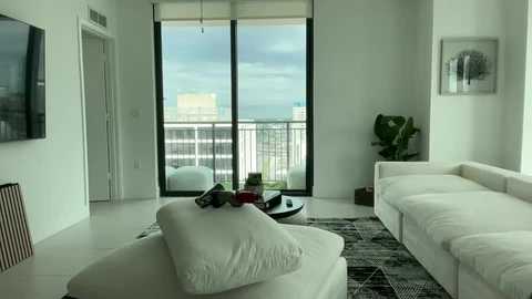 Video of a modern, minimalist living room with white furniture, including a sofa and armchair, and a large window revealing a cityscape. A potted plant and abstract art hang on the walls.