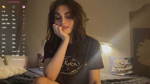 Video of a young woman with wavy dark hair, wearing a black T-shirt, resting her chin on her hand, in a dimly lit bedroom with a bed and wall art.