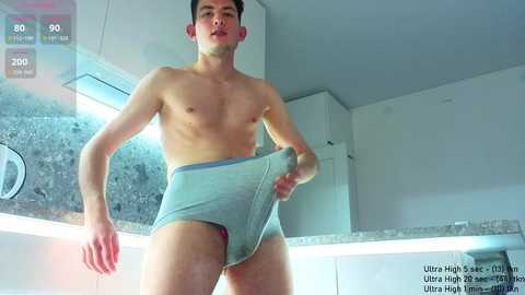 Media: Video of a fit, shirtless young man in light blue briefs, standing in a modern, white kitchen. He has a lean physique, fair skin, and dark hair. The background includes a granite countertop and stainless steel appliances.