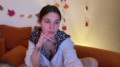 Video of a young woman with light skin, brown hair, and a tattooed hand, wearing a leopard-print hoodie, sitting on an orange sofa in a cozy room adorned with autumn-themed decorations.
