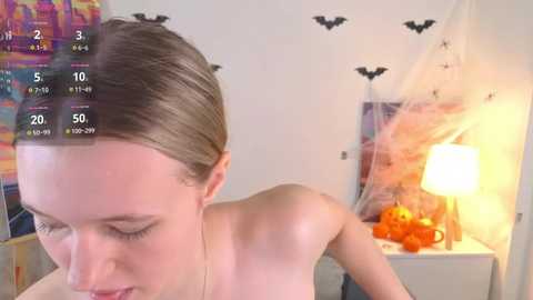 Video of a young woman with light skin and straight, light brown hair, partially undressed, in a brightly lit bedroom with Halloween decorations, including a bat garland, orange pumpkins, and a glowing lamp.