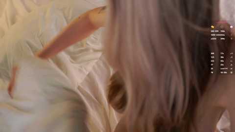 A video of a close-up, blurred face with long, wavy, light brown hair. The background features a white, wrinkled bedsheet. The image is soft-focused with a warm, golden hue.