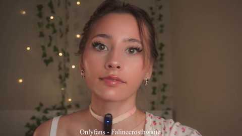 Video of a young Asian woman with light skin and brown hair in a casual setting, wearing a floral blouse and a choker. Background features fairy lights and green vines.