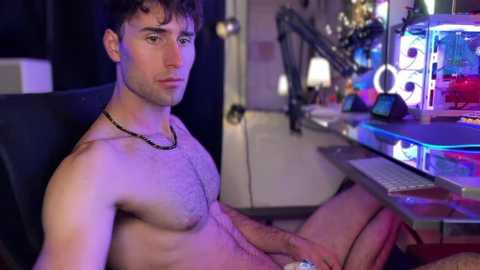 Video of a shirtless, fit, young white man with curly brown hair, wearing a black necklace, sitting in a chair in a dimly lit room with a gaming setup.