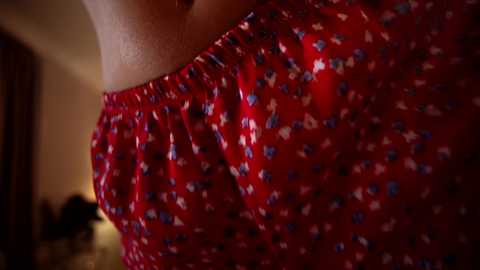 Close-up video of a woman wearing a red floral-patterned blouse with gathered neckline, in a dimly lit room with blurred background.