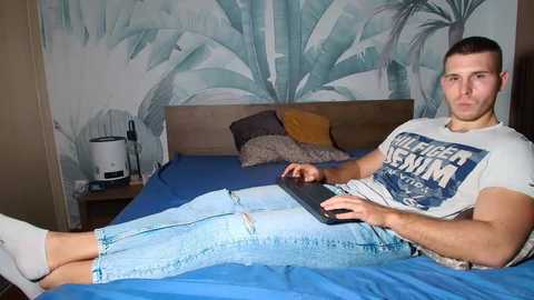 Video of a young man with short brown hair, wearing a white T-shirt with blue graphics and ripped jeans, lying on a blue bed, typing on a laptop, in a bedroom with a tropical wallpaper background.