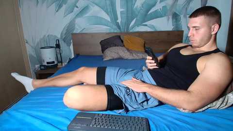 Video of a muscular, fair-skinned man with short brown hair lying on a blue bedspread, wearing a black tank top and blue shorts, playing with a smartphone. Background features a modern bedroom with a wooden headboard, a laptop, and a tropical mural.