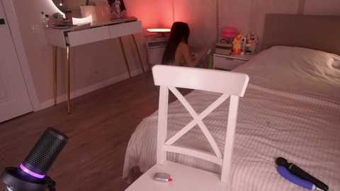 Video of a modern, dimly-lit bedroom with a white chair, white bed, and a woman sitting at a desk with a laptop.