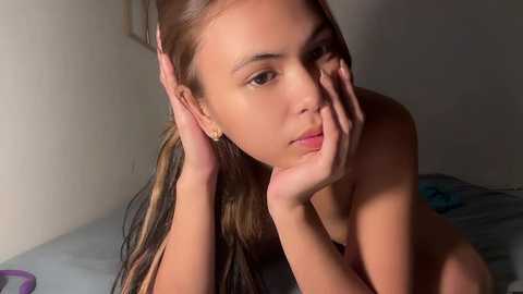 Media: Video of a young woman with light brown skin, long brown hair, and large brown eyes, posing topless on a bed with a grey blanket, hands resting on her face, in a dimly lit room.