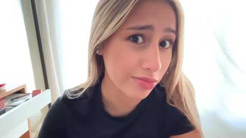 Video of a young Caucasian woman with fair skin and long, straight blonde hair. She has light makeup, brown eyes, and wears a black shirt. Background shows a window with white curtains, and a wooden table with a red book on it.
