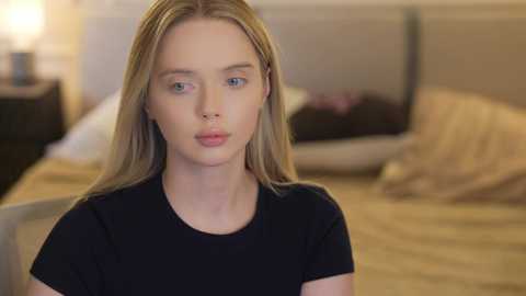 A video of a young Caucasian woman with light skin and long blonde hair, wearing a black T-shirt, sitting in a softly lit bedroom with beige bedding and pillows.