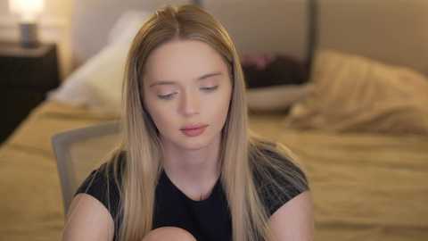 Video of a young Caucasian woman with long, straight blonde hair, wearing a black shirt, sitting on a bed in a softly lit bedroom.