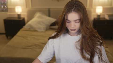 Media: A video of a young woman with long brown hair, wearing a white shirt, standing in a dimly lit bedroom with beige walls, two bedside tables, and lamps.