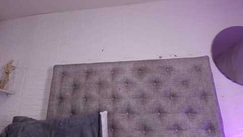 A video of a minimalist bedroom featuring a tufted, gray upholstered headboard against a white brick wall. A gray throw blanket is draped over a bed with a round mirror on the right side.