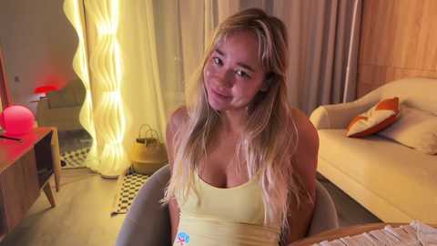 Video of a smiling, blonde, light-skinned woman in a yellow tank top, sitting on a modern chair in a cozy, dimly lit room with a checkered rug, wooden furniture, and a tall, wavy lamp.