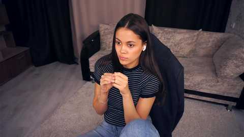 Video of a young woman with medium brown skin, long black hair, and a slender physique, wearing a striped shirt and jeans, sitting on a gaming chair in a cozy living room with beige couches and dark curtains.