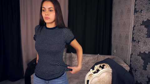 Video of a young woman with straight, dark hair, wearing a striped top, standing in a dimly lit, elegantly decorated room with floral-patterned walls and dark curtains.