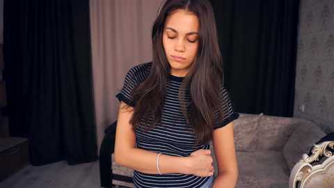 Video of a young woman with long dark hair, wearing a black and white striped shirt, standing in a dimly lit room with beige and dark curtains. She has a contemplative expression.