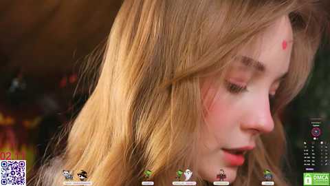 Video of a young Caucasian woman with long blonde hair, wearing a red heart on her forehead, in a close-up profile shot, set against a blurred background.