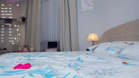 A video of a modern bedroom featuring a white bed with blue floral sheets, a pink toy on the bed, beige walls, a headboard, and a nightstand with a lamp.