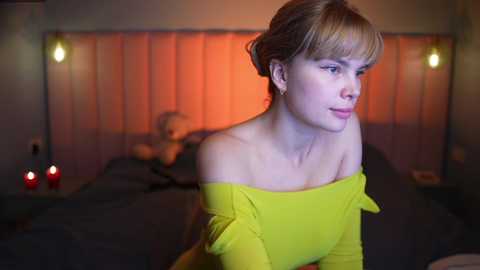 Video of a blonde woman in a bright yellow off-shoulder dress, leaning on a bed with red candles, in a dimly lit bedroom with orange curtains.