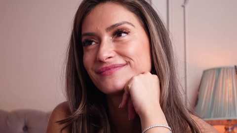 Video of a smiling, light-skinned woman with long, straight brown hair, resting her chin on her hand, wearing a light-colored top, against a softly lit, beige wall background.