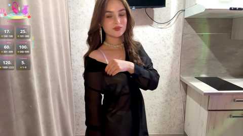 Video of a young woman with long brown hair, wearing a sheer black blouse and pearl necklace, in a modern kitchen with white marble countertops and appliances.