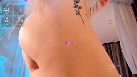 Video of a topless woman with light skin, visible tattoos, and a pink sticker on her back, taken indoors with a digital overlay showing a video game score.