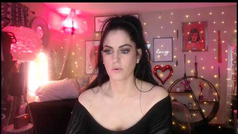 A video of a pale-skinned woman with long black hair, wearing black makeup and a black off-shoulder top, in a dimly lit room adorned with fairy lights, framed art, and a red heart-shaped mirror.