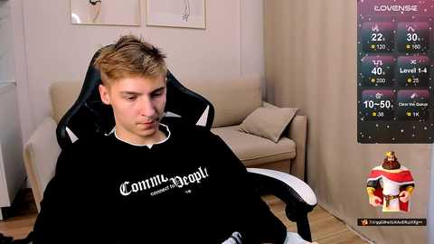 Video of a young man in a black gaming chair, wearing a black shirt with white text. A beige couch is in the background, with a colorful fitness chart on the wall.