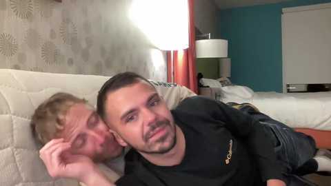 A video of two men, one sleeping on a white bed with a patterned sheet, and the other awake, smiling, with a black shirt and beard, in a modern bedroom with teal walls and a lamp.