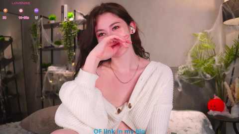 Video of a young woman with fair skin, dark hair, wearing a white cardigan and headphones, sitting on a bed with a green plant and a red pumpkin.