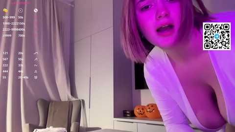 A video of a young Asian woman with shoulder-length, straight, pink hair, wearing a low-cut white top, revealing her small breasts. She's indoors, illuminated by purple lighting, with a QR code and camera overlay.