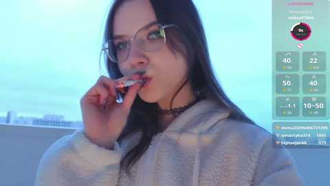 A video of a young woman with long black hair, wearing glasses and a gray robe, biting a lollipop, in a modern room with a digital temperature gauge.