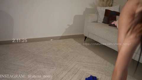 Video of a nude woman standing on a beige carpeted floor, facing a white couch with pillows, in a minimalist room with a cream-colored wall.