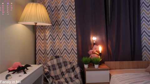 Video of a cozy bedroom with a white desk, a plaid chair, a wooden nightstand, a lamp, a flower arrangement, and chevron-patterned curtains.