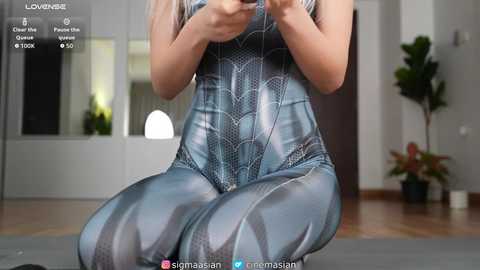 Media: Video of a light-skinned woman in a shiny, metallic blue Spider-Man suit kneeling, holding a smartphone, in a modern, minimalist living room with potted plants.