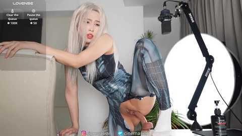 Media: Video of a slender, platinum blonde woman in a plaid dress, sitting provocatively on a white couch, with a large studio light in the background.