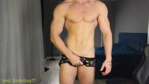 Video of a shirtless, muscular, fair-skinned man in black patterned underwear, standing in a dimly lit bedroom with a bed and gray curtains.