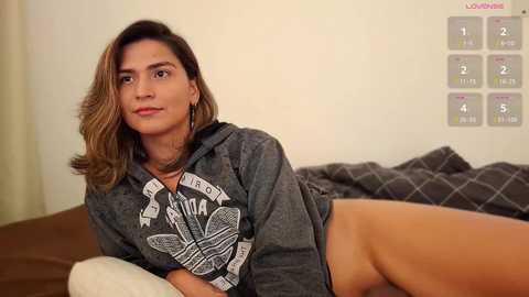 Video of a light-skinned woman with medium-length, wavy brown hair, wearing a grey hoodie, lying on a bed with a brown and grey blanket, in a cozy bedroom with a wall chart of elements.