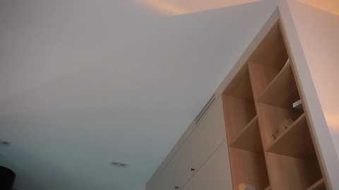 Media: A video of a minimalist, modern interior featuring a geometrically shaped white shelving unit with light wooden inserts, against a cloudy, pale blue sky.